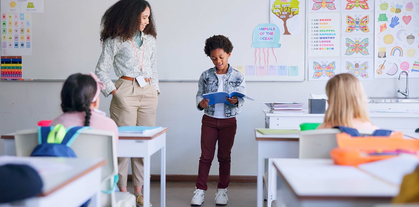 A young boy presents in front of his class with a female teacher standing beside him, in a colorful elementary school classroom.