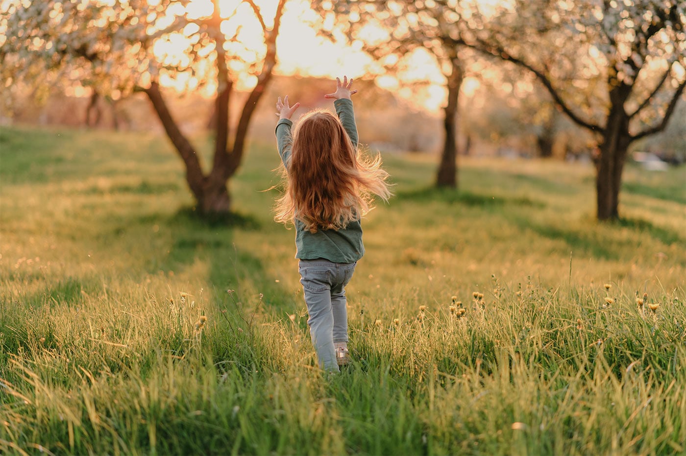 Young girl with flowing hair running joyfully in a sunlit orchard, casting a warm glow on the grass and blooming trees.