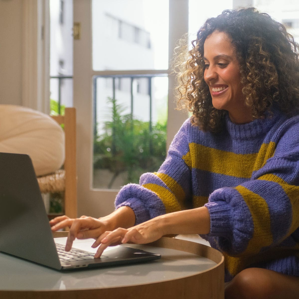 A person in a purple and yellow striped sweater is typing on a laptop at a small table, with a bright, sunny window in the background.