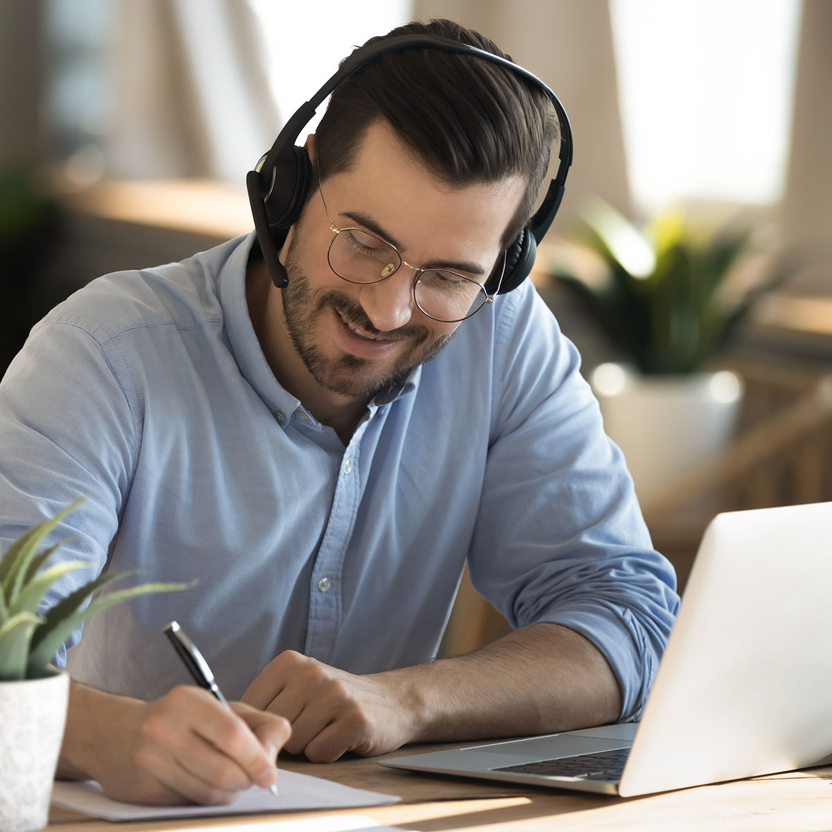 Smiling young Caucasian man in headphones glasses sit at desk work on laptop making notes. Happy millennial male in earphones watch webinar or training course or computer, study online from home.