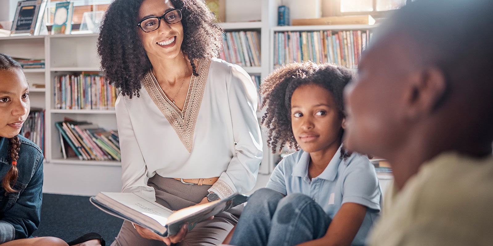 A teacher reads a book to three attentive children in a brightly lit library filled with books.