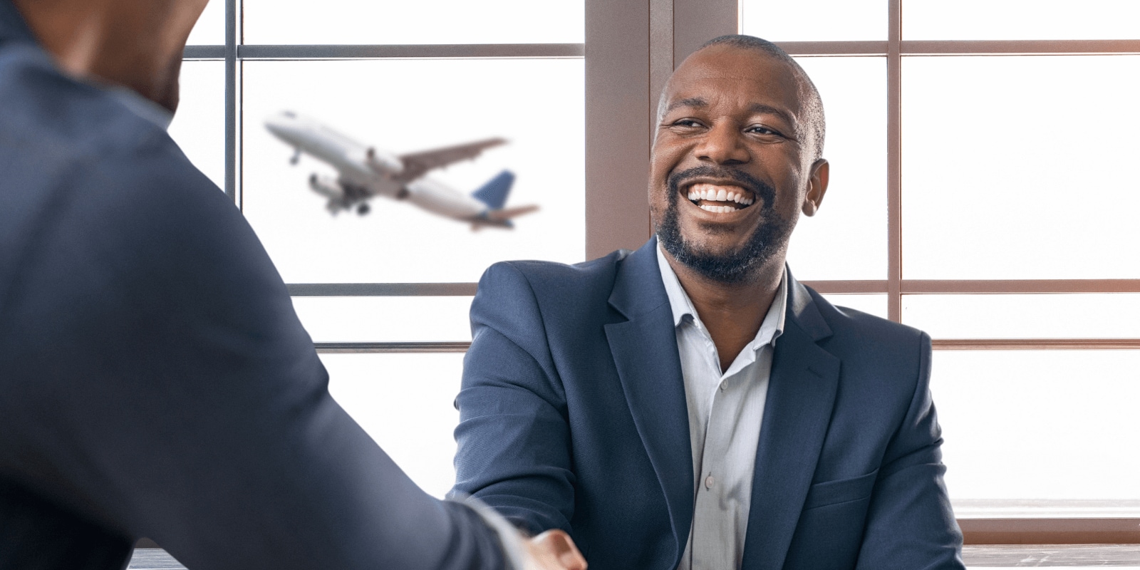 Two businessmen in suits sit at a table near a large window with a plane flying outside.