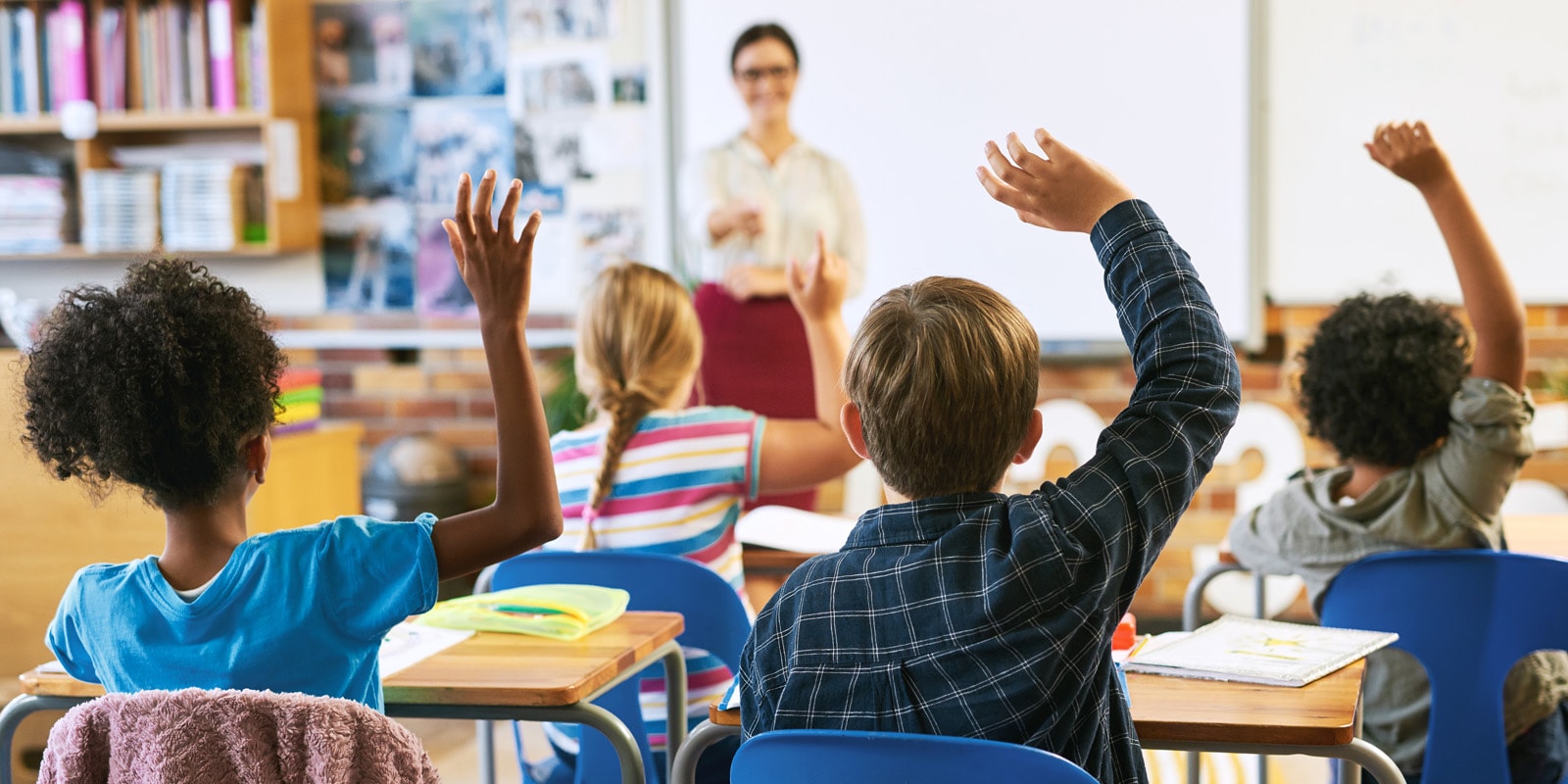 Children in a classroom raising their hands eagerly as a teacher stands in the background