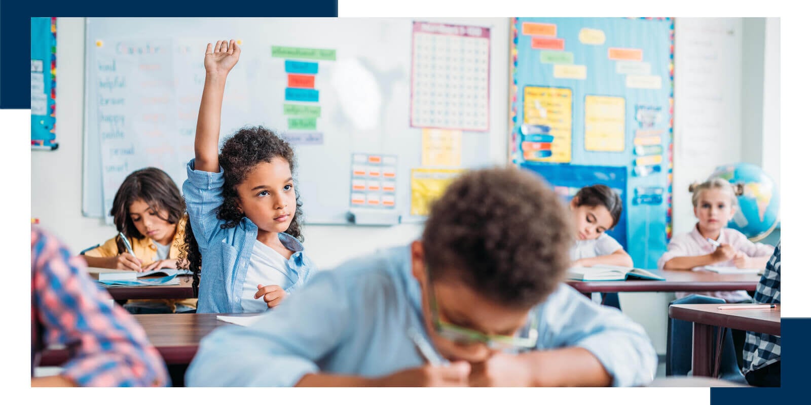 Classroom with students at desks, one raising hand; colorful educational posters on the wall.