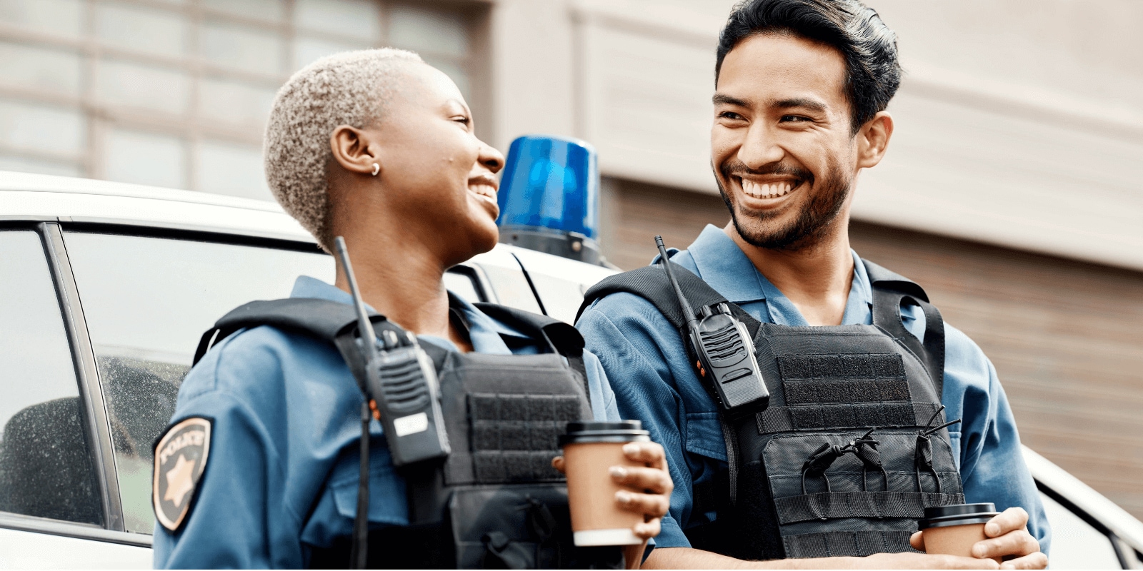 Two police officers in bulletproof vests stand beside a patrol car, one holding a coffee cup, both equipped with radios and badges.